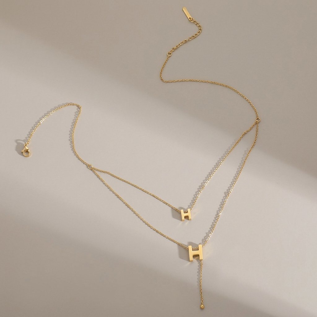 Double-layer gold H necklace with two H pendants and a chain tail displayed on a warm-gray background.