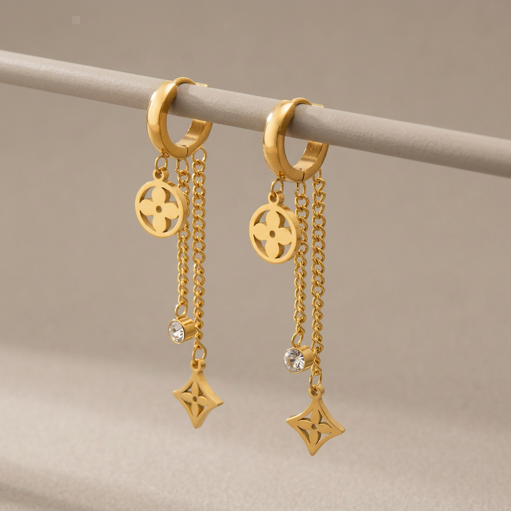 Gold clover dangle hoop earrings with chain drops and clear stones hanging from a warm-gray support.