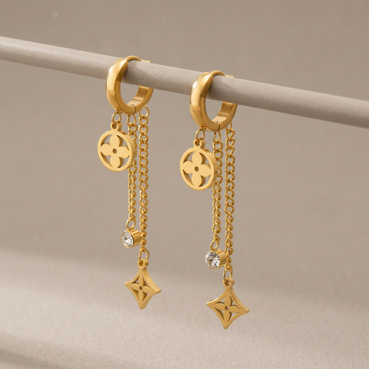 Gold clover dangle hoop earrings with chain drops and clear stones hanging from a warm-gray support.