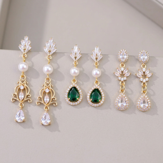 Three pairs of gold bridal earrings with pearl filigree, green teardrop and clear leaf teardrop designs on a warm gray background