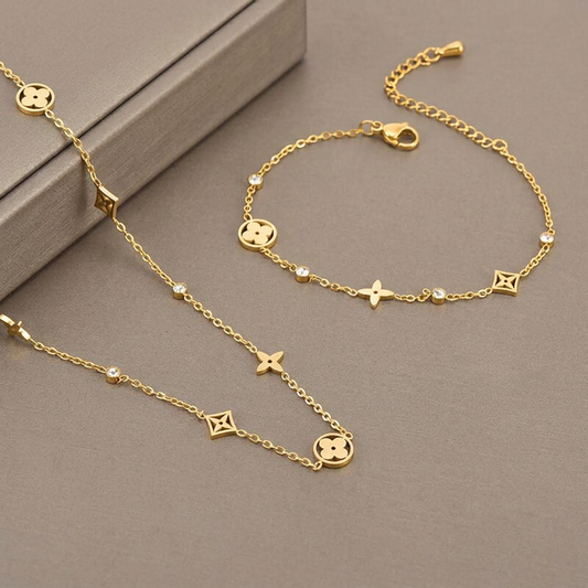 Gold clover necklace and matching bracelet displayed on a warm-gray studio background.