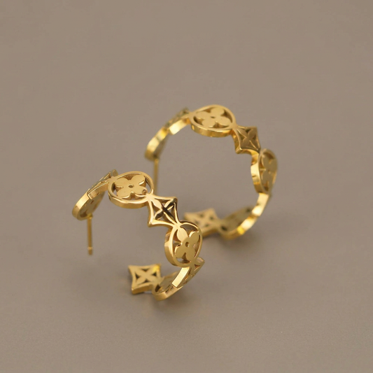Pair of gold clover hoop earrings on a warm-gray studio background.