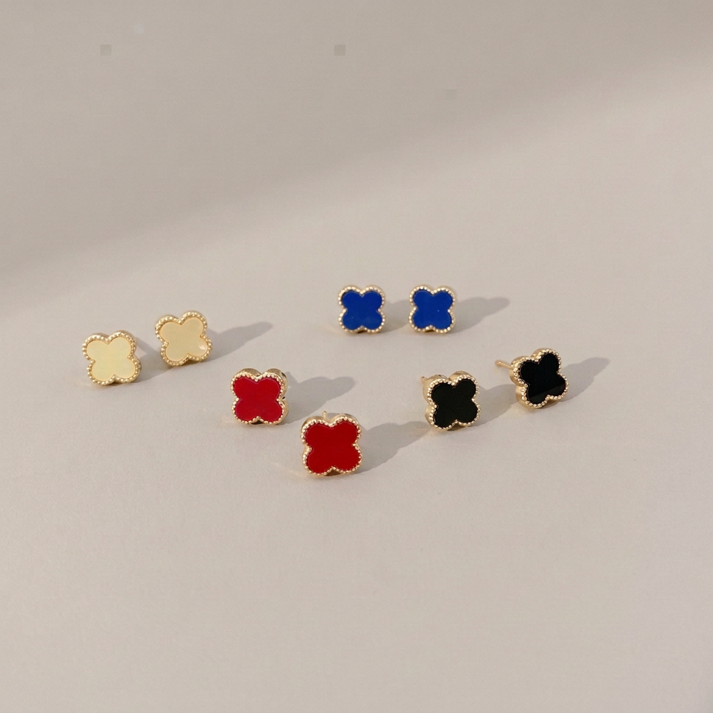 Four pairs of gold-tone clover stud earrings in pale cream yellow, blue, red and black on a warm gray background