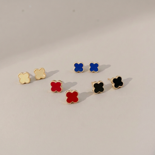 Four pairs of gold-tone clover stud earrings in pale cream yellow, blue, red and black on a warm gray background